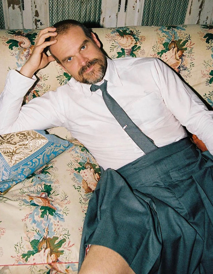 David Harbour seated casually on a floral couch wearing a white shirt, tie, and dark pleated skirt, looking thoughtful.