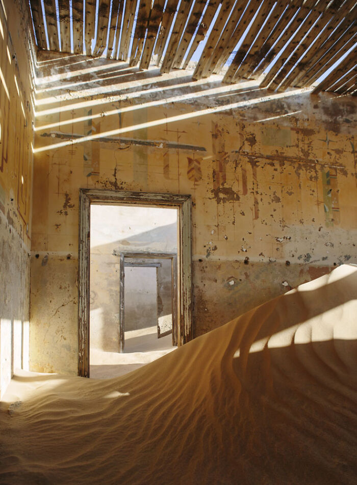 Abandoned home interior partially buried in desert sand with sunlight streaming through a broken roof structure.