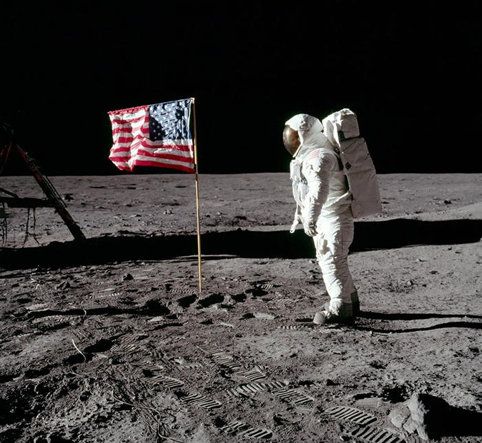 Astronaut standing on the moon next to the American flag with lunar surface and dark space in the background.