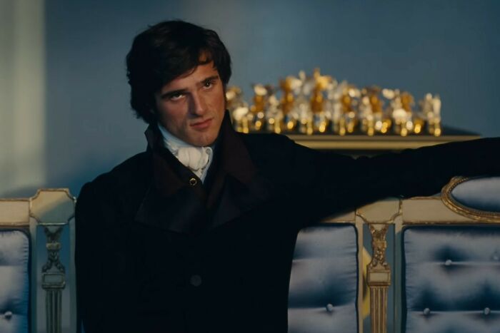 Young man in dark period costume sitting on ornate blue and gold settee in a scene from Wuthering Heights trailer.