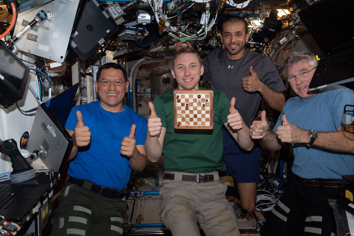 Four astronauts in a spacecraft give thumbs up around a floating chessboard, showcasing epic astronomy photos of space life.