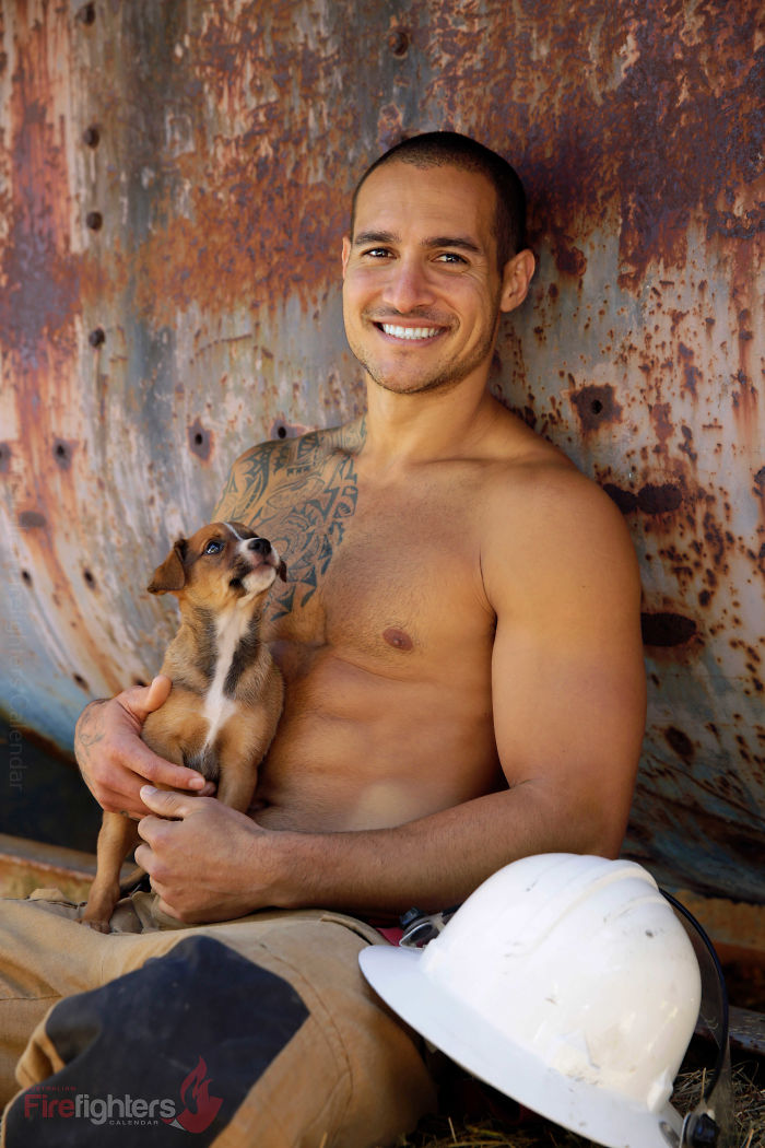 Smiling shirtless firefighter holding an adorable dog, sitting by a rusted wall with a white helmet nearby.