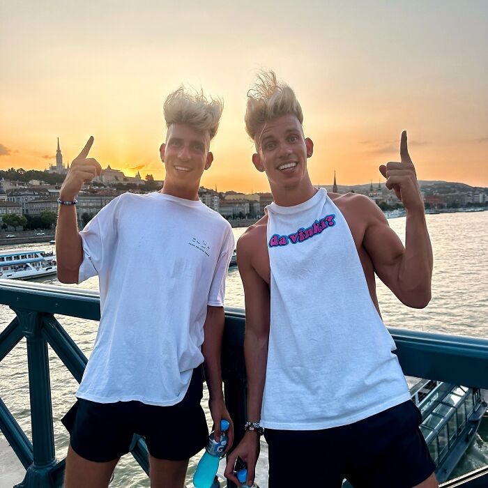 Twin celebrity brothers smiling and posing by a waterfront at sunset, showcasing celebrity twins enjoying a scenic view.