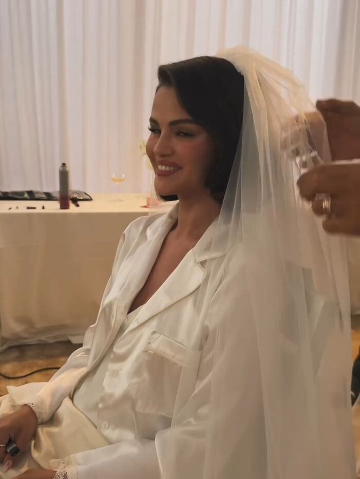Selena Gomez smiling while wearing a white bridal robe and veil, with hands adjusting the veil during wedding preparations.