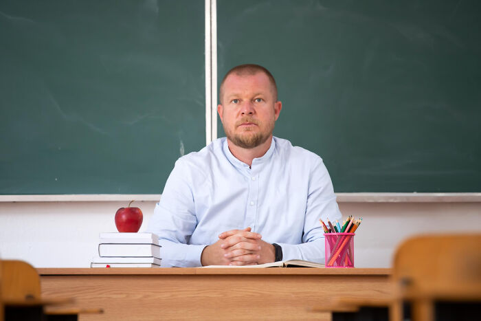 Man sitting at a classroom desk with books, an apple, and colored pencils in front of a chalkboard, looking serious and creepy.