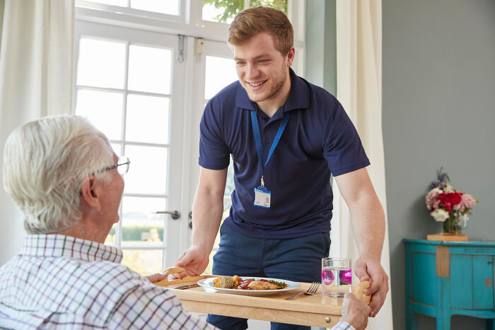 Caregiver serving lunch to elderly patient with dementia in a bright room, capturing unfiltered and funny moments.