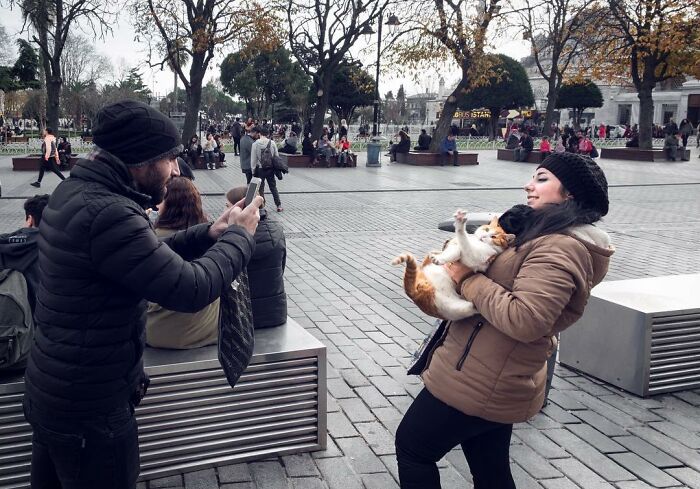 Man taking photo of woman holding cat on street in Istanbul, capturing everyday life with cats in urban setting.