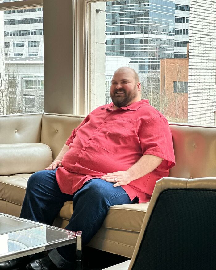 Plus-size activist wearing a red shirt sitting on a beige couch in an urban setting with skyscrapers outside. Plus-size activist wearing a red shirt sitting on a beige couch in an urban setting with skyscrapers outside.