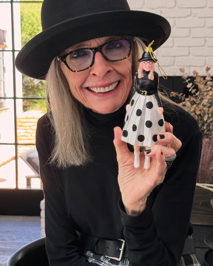 Diane Keaton smiling indoors, wearing a black hat and glasses, holding a doll dressed in similar black and white attire.