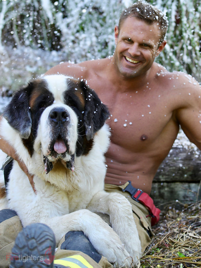 Firefighter sitting shirtless behind a large dog, both outdoors with water droplets falling around them.