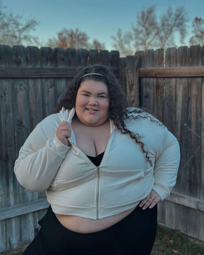 Plus-size activist in a cream jacket and black top posing outdoors by a wooden fence with a confident smile. Plus-size activist in a cream jacket and black top posing outdoors by a wooden fence with a confident smile.