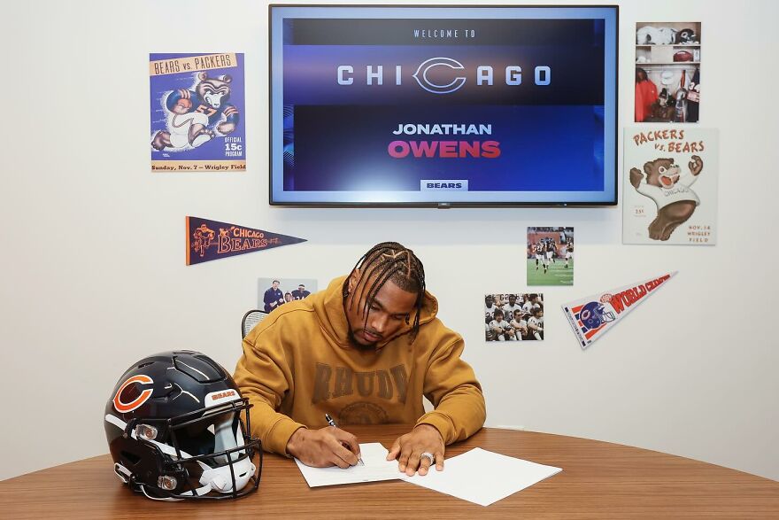 Jonathan Owens in a Chicago Bears hoodie signing contract with helmet and team posters visible in sports-themed room. Jonathan Owens in a Chicago Bears hoodie signing contract with helmet and team posters visible in sports-themed room.