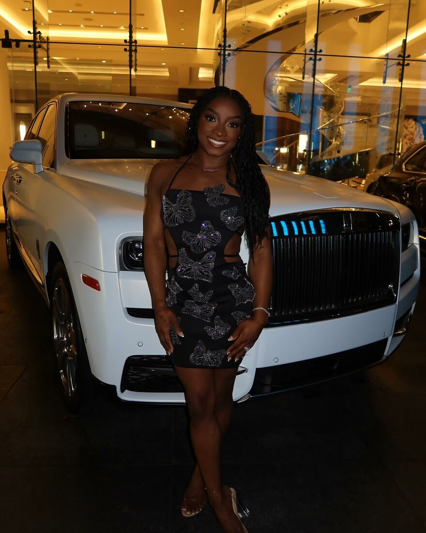 Simone Biles wearing a black butterfly dress standing in front of a white luxury car indoors, showcasing power couple net worth. Simone Biles wearing a black butterfly dress standing in front of a white luxury car indoors, showcasing power couple net worth.