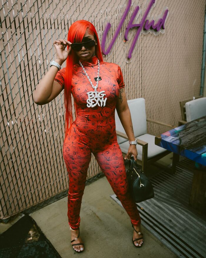 Rapper in red floral outfit and sunglasses posing indoors with a black handbag, linked to red hat allegations and twerking controversy.