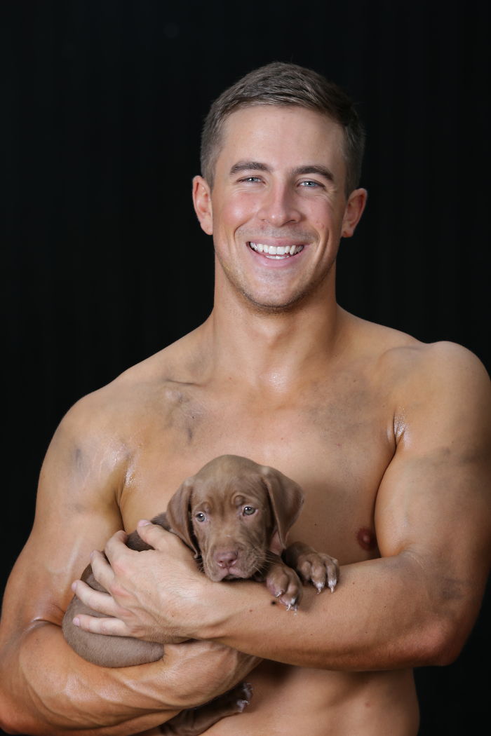 Shirtless firefighter with dirt marks smiling and holding an adorable brown puppy against a black background.