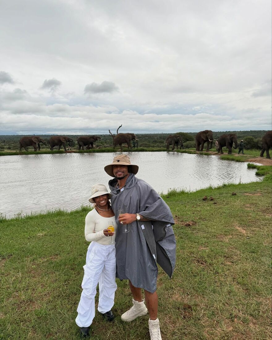 Jonathan Owens and Simone Biles smiling near a watering hole with elephants in the background on a cloudy day. Jonathan Owens and Simone Biles smiling near a watering hole with elephants in the background on a cloudy day.