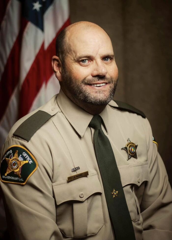 Police chief in uniform posing for official portrait with American flag in the background after leaked video controversy.