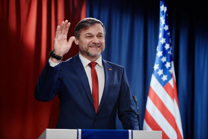 Man in suit waving during speech with American flag backdrop, capturing candid moments of a patient with dementia.