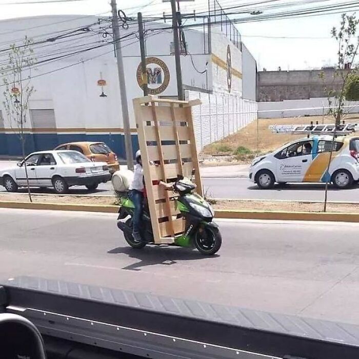 Person transporting a large wooden pallet on a scooter on a city street, a strange image that might need a priest or exorcist.