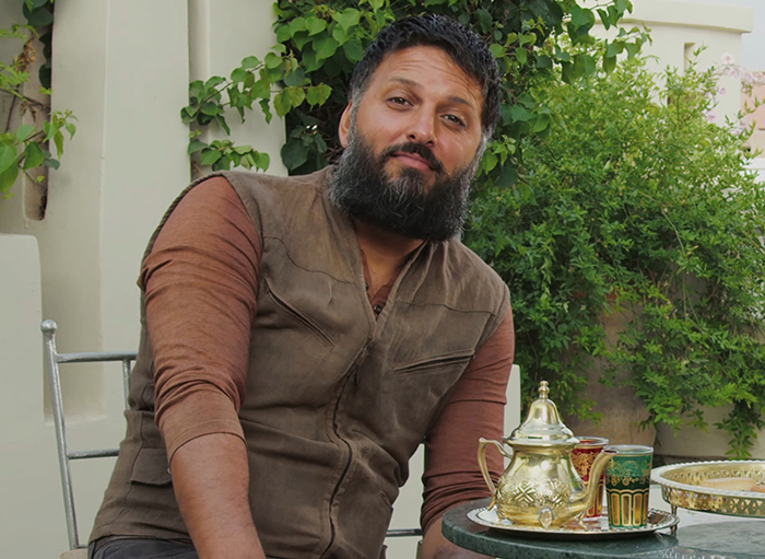 Man with a beard and brown vest sitting outdoors at a table with a golden teapot and colorful cups, greenery behind.