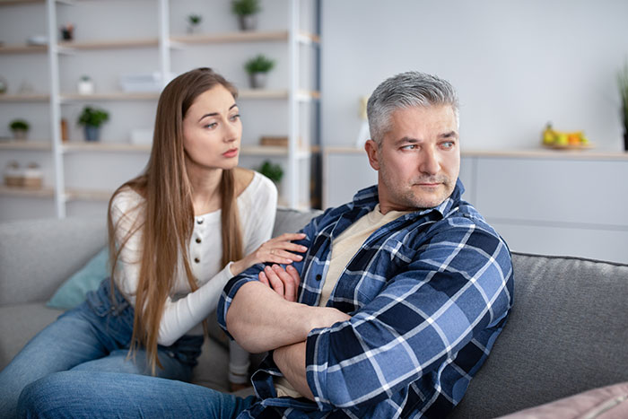 Woman looking concerned while talking to boyfriend&rsquo;s grown son sitting with arms crossed on a couch at home
