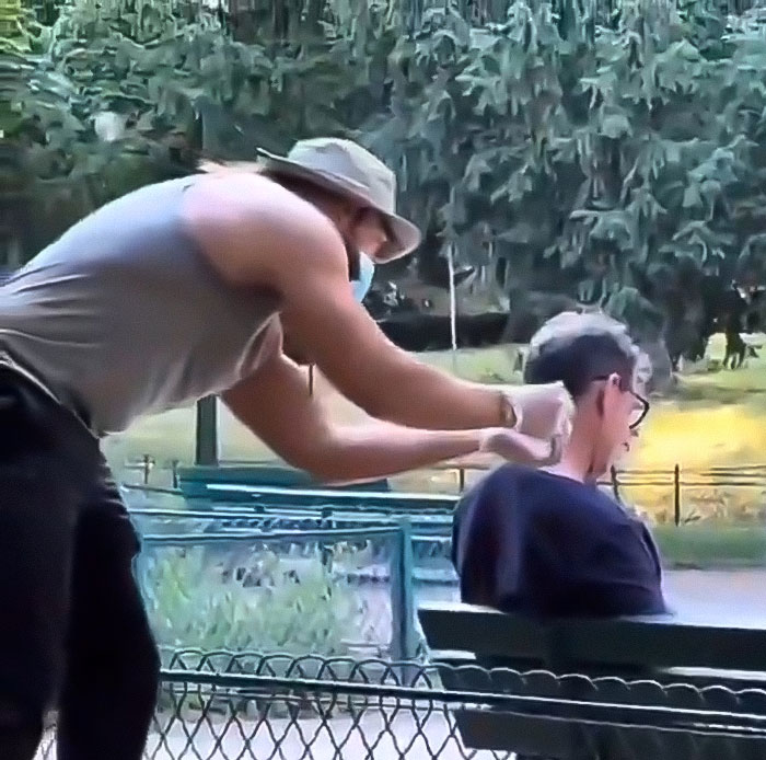 Man wearing a hat and mask pretending to perform a fake syringe attack prank on a seated young man in a park.