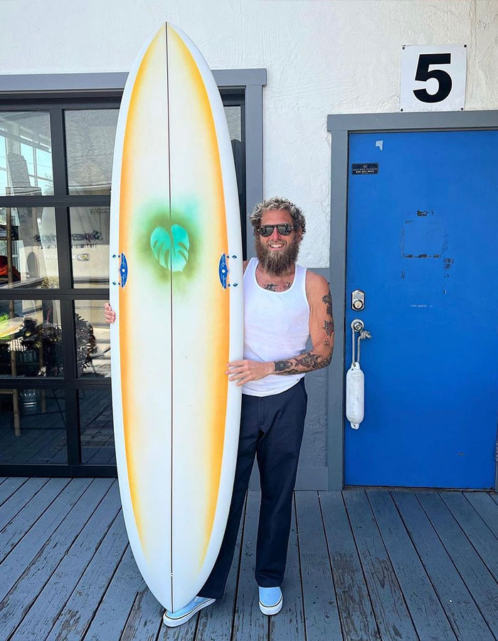 Jonah Hill showing major transformation, wearing sunglasses and tank top, standing with surfboard by a blue door outdoors.