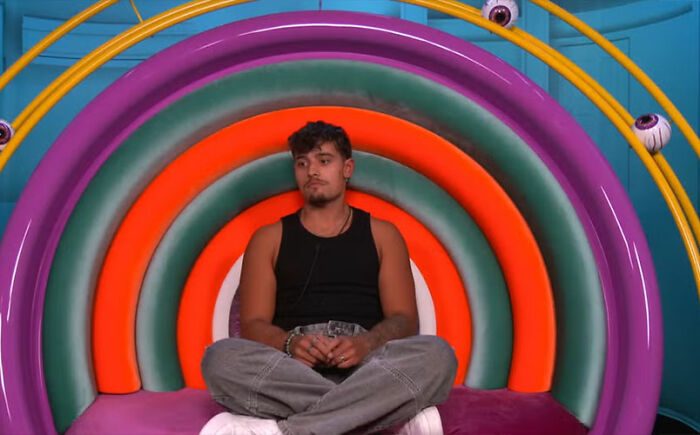 Big Brother housemate sitting against colorful circular wall, fans furious and calling for immediate exit after tense moment.