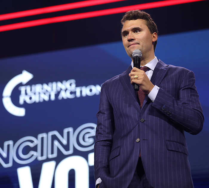 Charlie Kirk speaking at Turning Point Action event, wearing a striped suit and holding a microphone on stage.