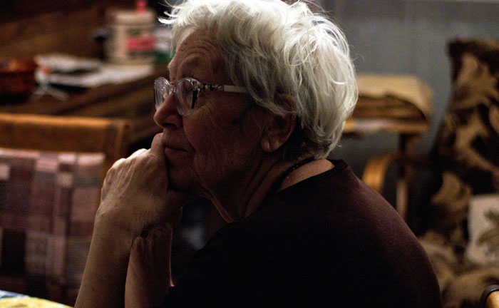 Elderly woman with glasses sitting thoughtfully indoors, evoking stories daycare workers heard about family secrets.