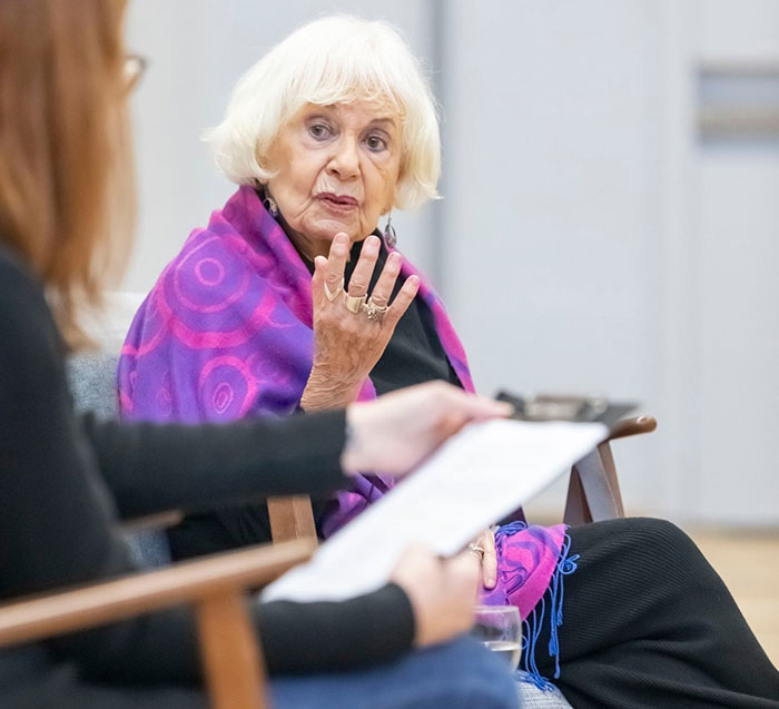 Holocaust survivor Ruth Posner speaking during an event, wearing a purple shawl and engaging with a seated person.