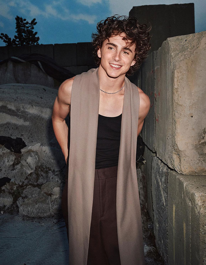 Timoth&eacute;e Chalamet smiling outdoors at dusk wearing a sleeveless top and long beige scarf with curly hair.
