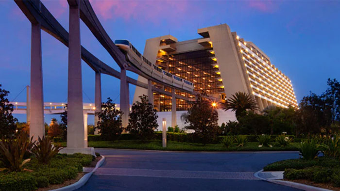 Monorail passing in front of a brightly lit Disney resort building at dusk, surrounded by trees and landscaping.