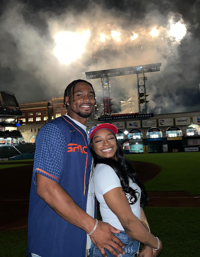 Simone Biles and Jonathan Owens smiling together at a stadium with fireworks and night sky in the background.