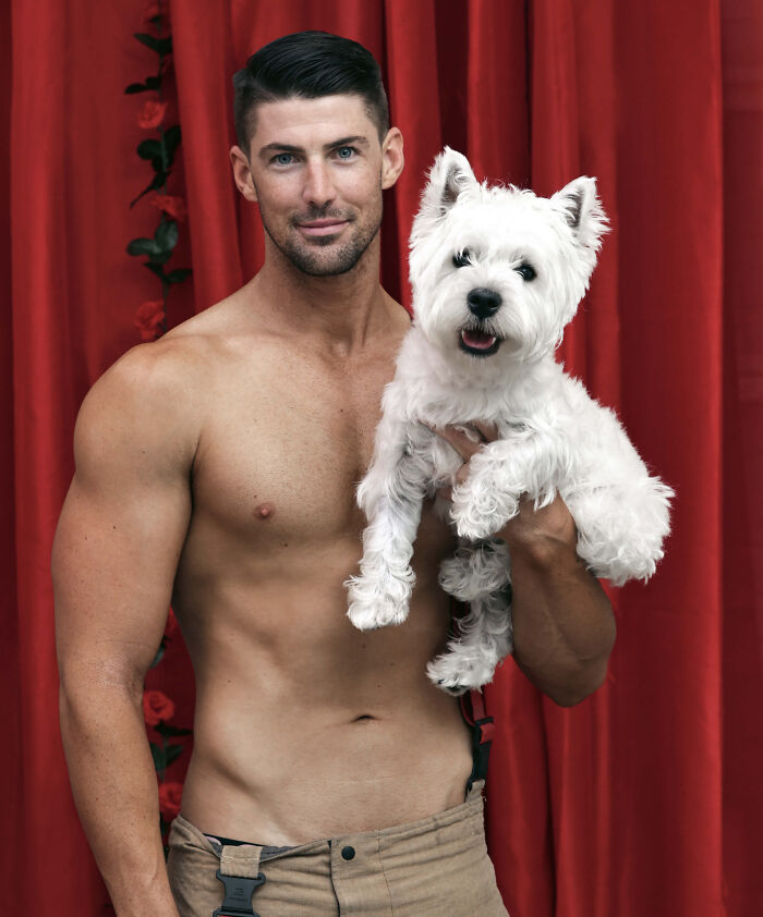 Shirtless heroic firefighter holding an adorable white dog in front of a red curtain for an Australian calendar photo.
