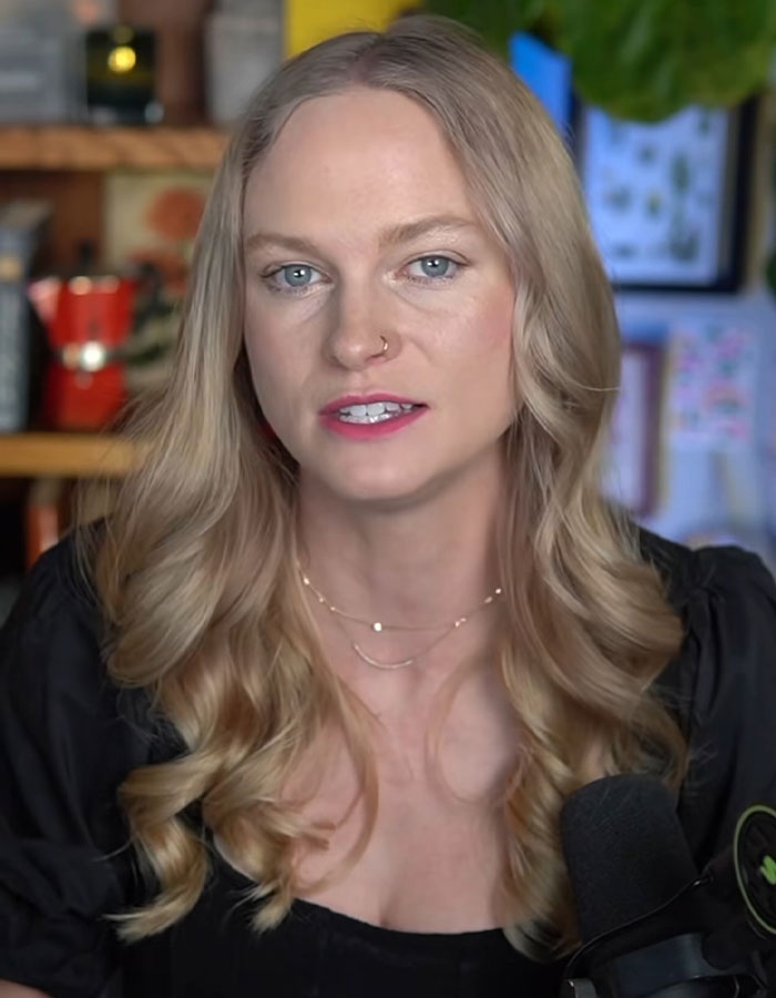 Young woman with long blonde hair discussing Mormon Backflip Theory trend in a casual home recording setup with microphone.