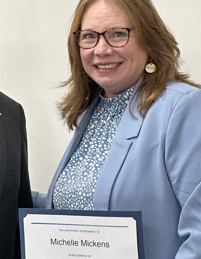 Teacher of the year finalist Michelle Mickens holding certificate, smiling in professional attire indoors.