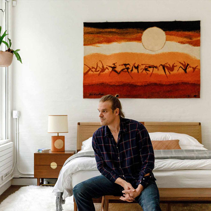 Man sitting on a bed in a modern bedroom, with a large colorful tapestry on the wall, reflecting on Lily Allen's album.