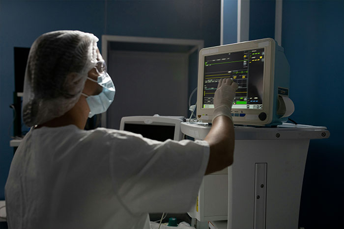 Medical professional in protective gear monitoring vital signs on a screen during post cosmetic surgery care in a dimly lit room.