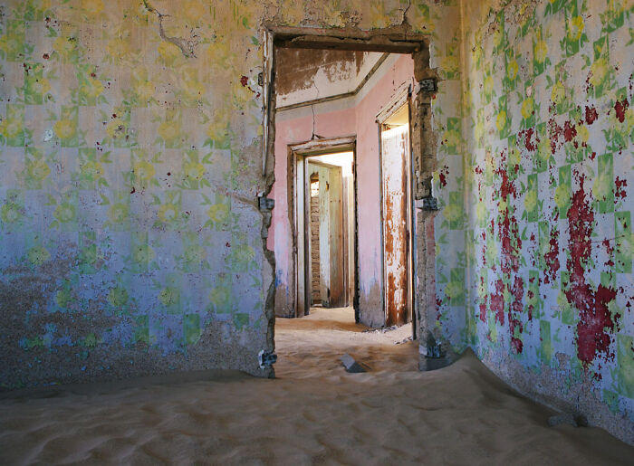 Abandoned home interior with sand partially filling rooms and peeling floral wallpaper in desert setting.