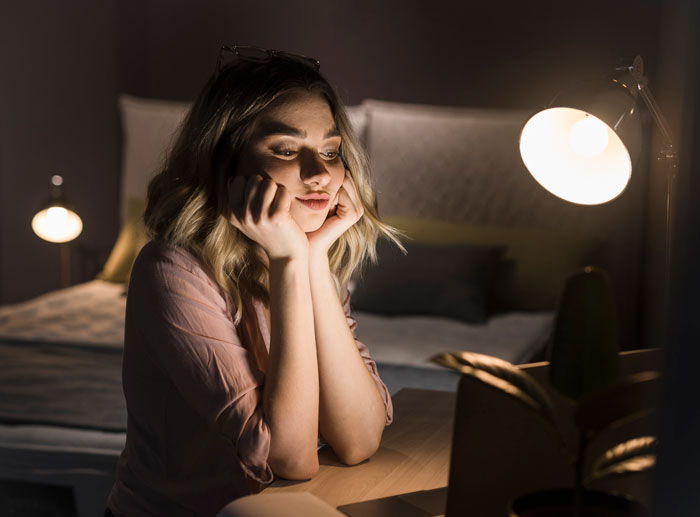 Young woman sitting at a desk in a dimly lit room, reflecting quietly on experiences with narcissistic mothers.