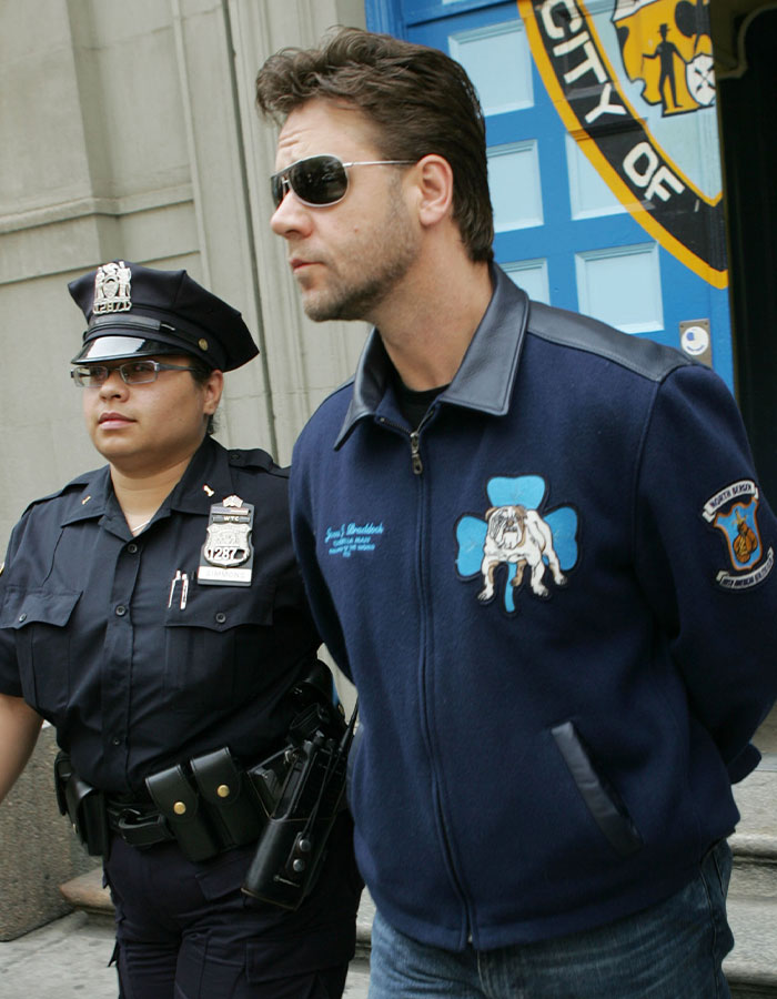 Police officer escorting a man wearing sunglasses and a blue jacket with a bulldog patch outside a building entrance.