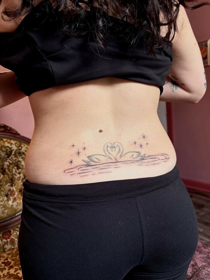 Lower back tattoo of two swans forming a heart shape surrounded by stars, seen on a person wearing black clothing.