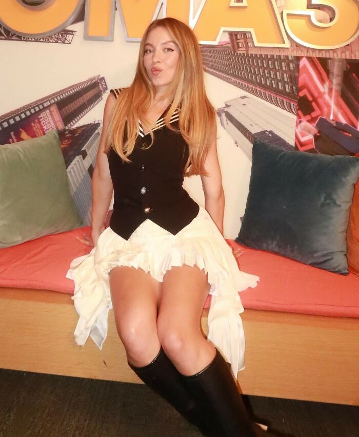 Woman with long hair sitting indoors wearing a black vest and white skirt, related to rapper red hat allegations topic.