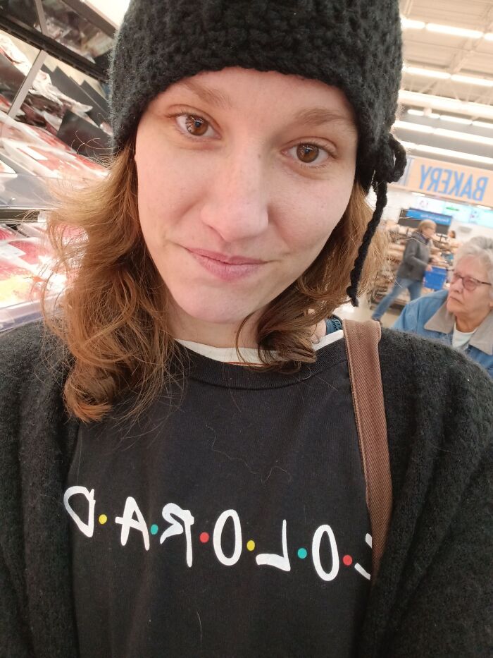 Woman in a black hat and sweater smiling in a store, with a cooler and a background showing bakery and other shoppers.