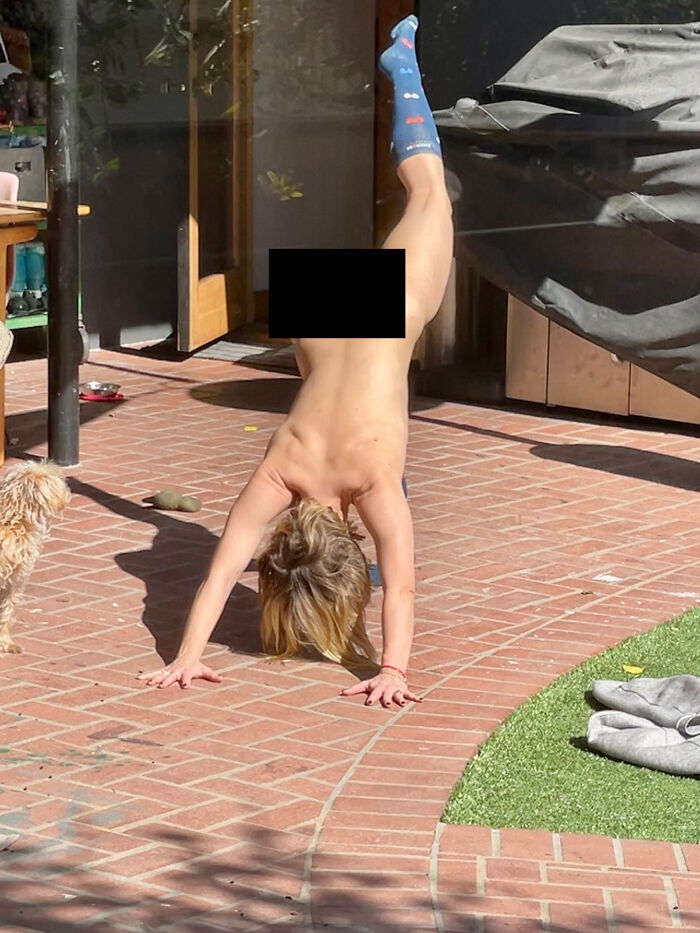 Kristen Bell doing a handstand outside on brick patio with one leg raised while a small dog looks on nearby.