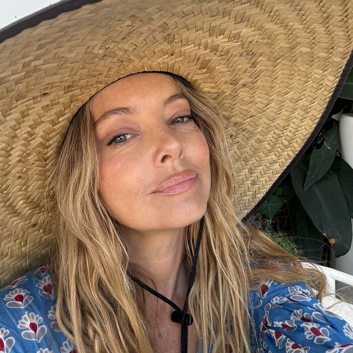 Bijou Phillips wearing a large straw hat and floral top, posing outdoors with natural makeup and loose wavy hair.