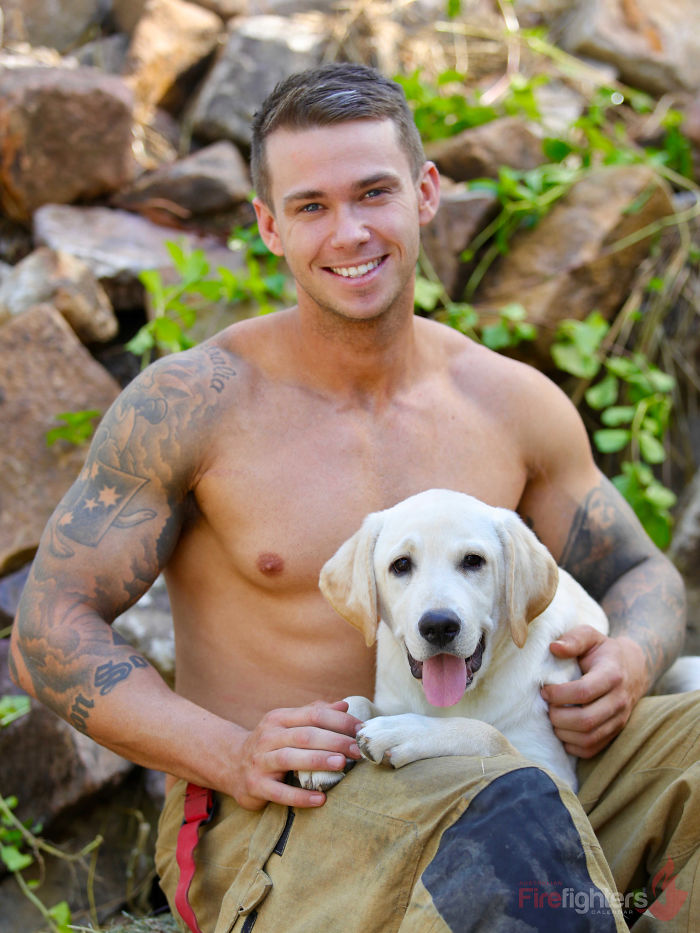 Shirtless Australian firefighter with tattoos sitting outdoors, smiling and holding an adorable white Labrador dog.