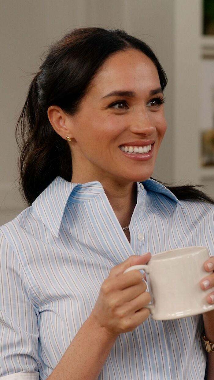 Meghan Markle smiling and holding a white mug during Paris Fashion Week appearance with casual striped shirt.