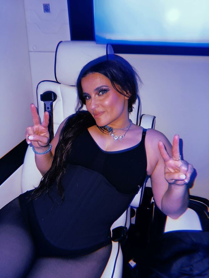 Nelly Furtado seated in a chair, wearing black outfit and jewelry, flashing peace signs with both hands in a relaxed pose.